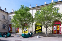 Toourism exhibition graphic design by LWZ Studio displayed on the exterior windows of the Architekturzentrum Wien Az W courtyard, featuring bold yellow and red typography with green outdoor seating. LWZ Design and Animation Vienna