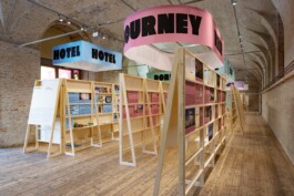 Spacious exhibition design for Toourism at Architekturzentrum Wien by LWZ Studio, featuring modular wooden stands and large circular hanging banners reading HOTEL and JOURNEY. LWZ Design and Animation Vienna