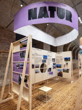 Interior view of the Toourism exhibition graphic design at Architekturzentrum Wien, showing wooden display structures with headphones and a large lilac hanging banner reading NATUR. LWZ Design and Animation Vienna