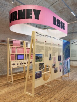 Toourism exhibition graphic design and spatial layout by LWZ Studio at Architekturzentrum Wien, showcasing a large suspended pink oval banner with the text JOURNEY and REISE over wooden information displays. LWZ Design and Animation Vienna