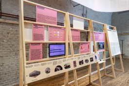 Detail view of the Toourism exhibition graphic design by LWZ Studio, highlighting pink infographic panels and an embedded digital screen mounted on a light wooden display stand against a brick wall. LWZ Design and Animation Vienna