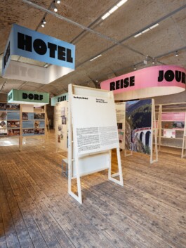 A wide view of the comprehensive Toourism exhibition graphic design by LWZ Studio, featuring wooden structures, informative panels, and colourful overhead banners reading Hotel, Dorf, Stadt, and Reise. LWZ Design and Animation Vienna