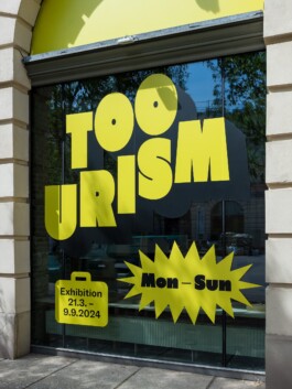 A close-up view of the Toourism exhibition graphic design by LWZ Studio, featuring bold yellow typography vinyl stickers on a large glass window, showing the main title, exhibition dates, and an opening hours starburst element reading Mon-Sun. LWZ Design and Animation Vienna