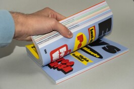 A close-up view of a hand flipping through the exhibition catalog designed by LWZ Studio and Manuel Radde for the Toourism exhibition, showcasing the colorful shapes and bold pop-art style page layouts. LWZ Design and Animation Vienna
